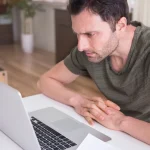 Hombre con gesto de impaciencia mirando la pantalla de su portátil mientras la página web tarda en cargar